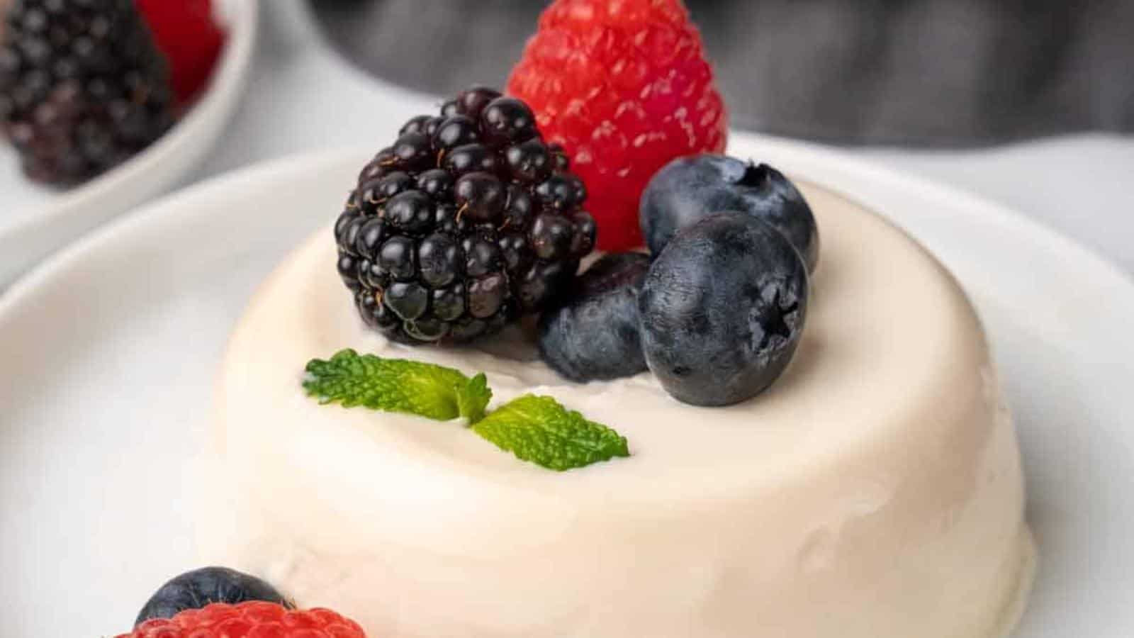 A creamy panna cotta sits on a white plate, topped with fresh blackberries, raspberries, blueberries, and mint leaves. The silky texture contrasts with the vibrant, colorful berries.