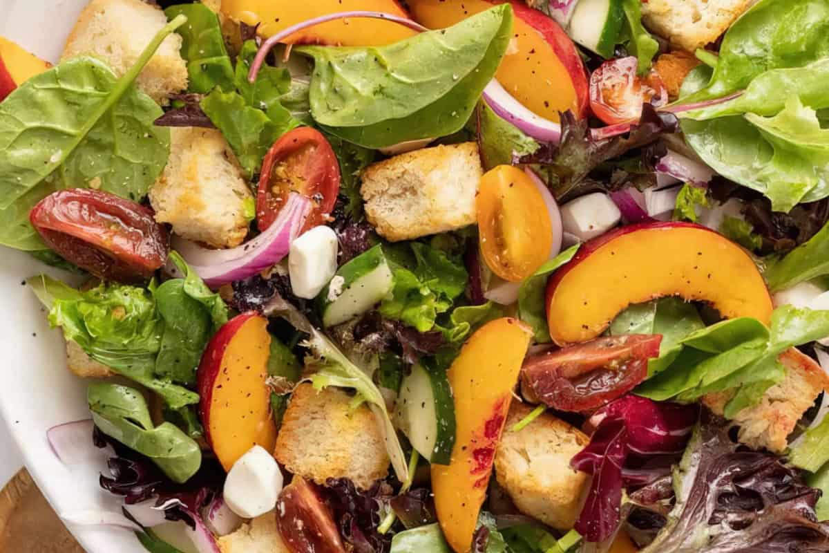 A vibrant salad featuring fresh greens, sliced peaches, cherry tomatoes, red onion, croutons, and small cubes of cheese. The ingredients are mixed together, creating a colorful and appetizing appearance.