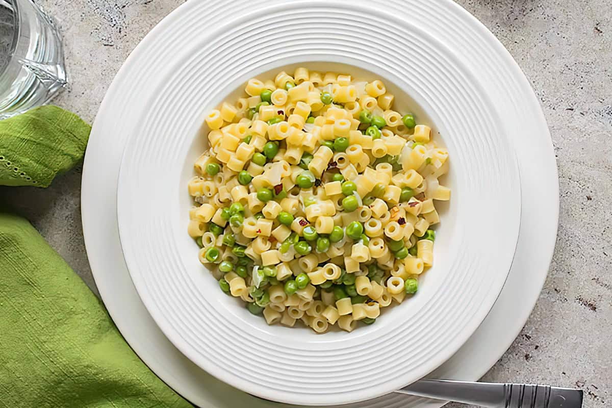 A white plate of pasta topped with peas and sprinkled with red pepper flakes. A green napkin and a fork are placed beside the plate, on a white surface.