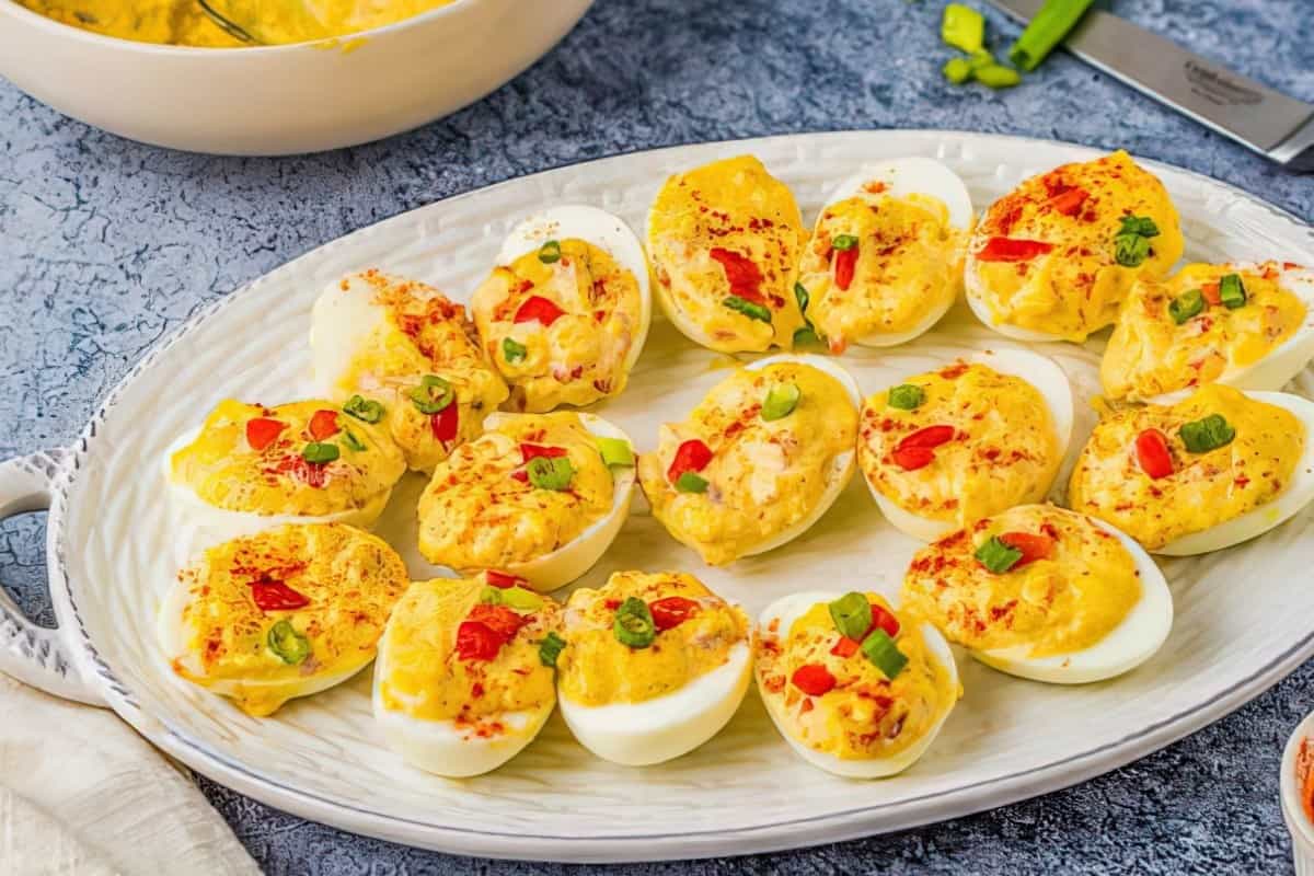 A platter of deviled eggs topped with paprika and sliced green onions, garnished with small red pepper pieces, on a textured blue surface. A serving bowl is partially visible in the background.