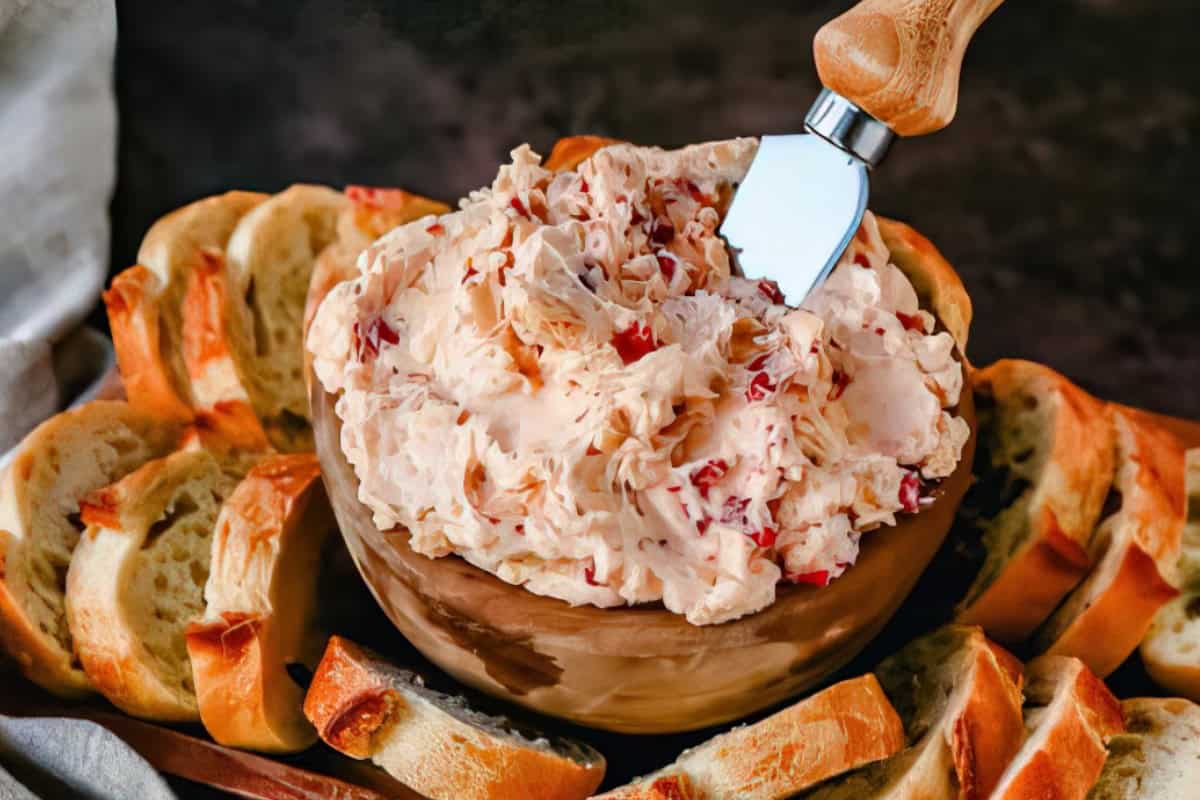 A wooden bowl filled with a creamy spread containing chopped ingredients, placed on a platter surrounded by slices of toasted bread. A spreading knife with a wooden handle is inserted into the spread.