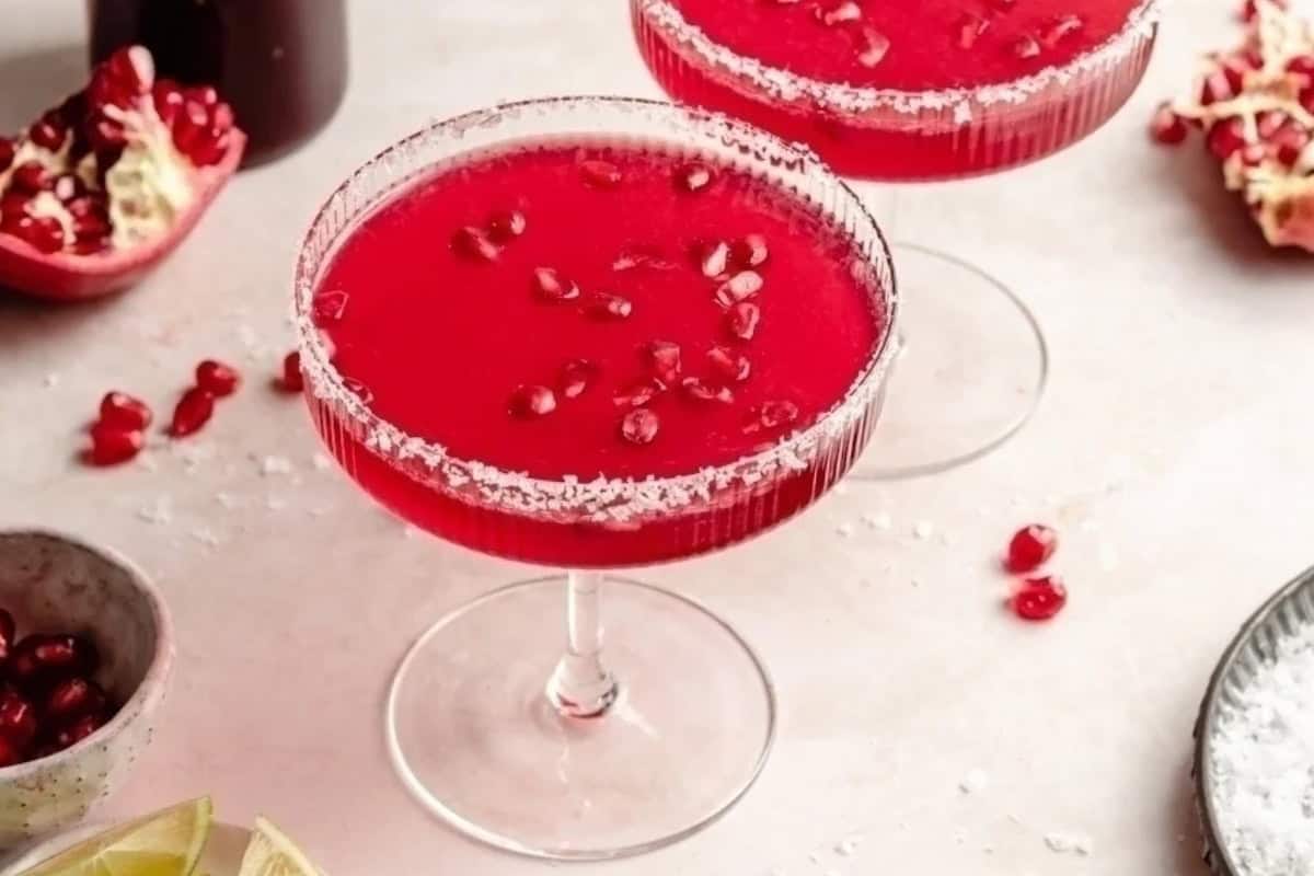 Two glasses of vibrant red pomegranate margaritas with salted rims are garnished with pomegranate seeds. Nearby, a halved pomegranate and lime slices are visible on a light-colored surface.