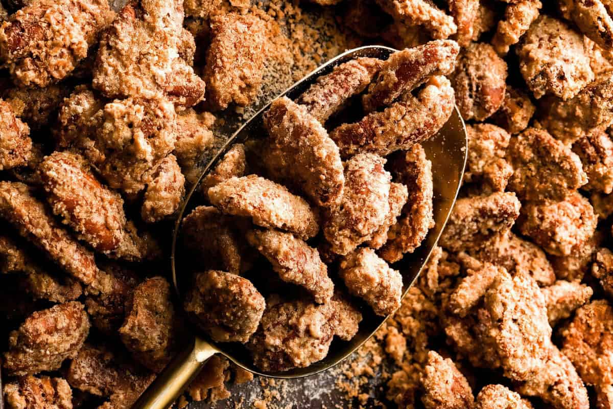A close-up of a spoon scooping sugar-coated pecans. The pecans are covered in a crumbly, cinnamon-spiced mixture and are scattered across a baking sheet. The image shows the rich texture and crunchiness of the snack.