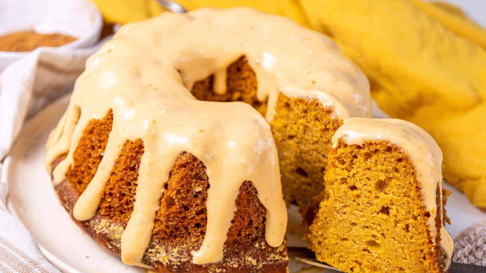 A glazed bundt cake with a slice cut out. A yellow cloth and a bowl in the background.
