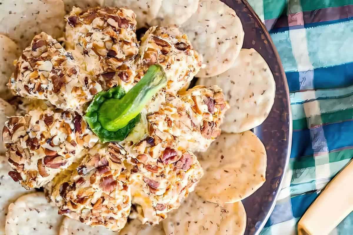 A cheese ball coated with chopped nuts, shaped like a pumpkin, is garnished with a green pepper stem. It is surrounded by round crackers on a plate, against a colorful striped tablecloth.