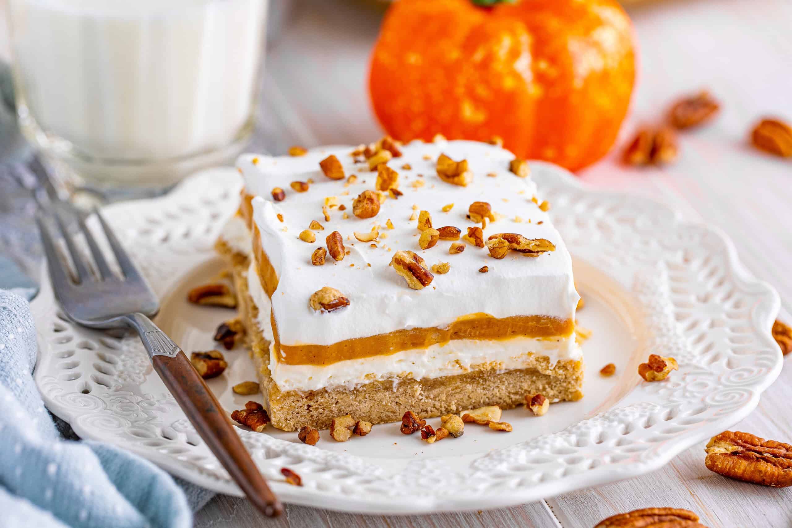 The image features a slice of layered pumpkin dessert topped with whipped cream and pecans, placed on a white plate. Beside the plate are a fork and a small pumpkin.