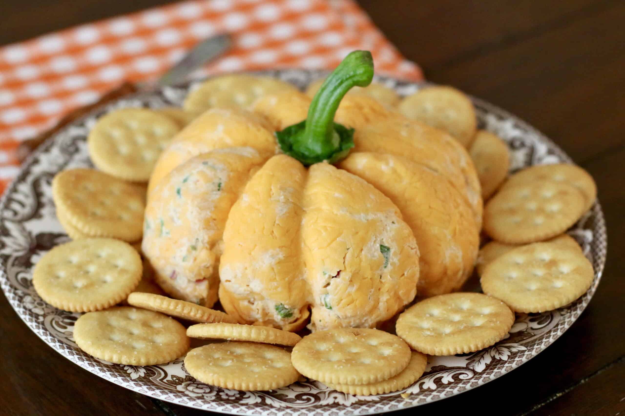 A cheese ball shaped like a pumpkin sits on a patterned plate, with round crackers encircling it. An orange checkered napkin is visible in the background.