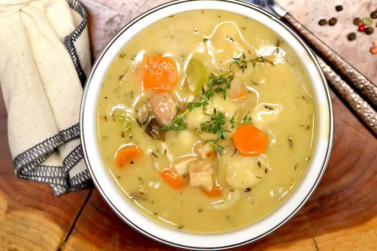 A bowl of soup with carrots, celery, and herbs on a wooden table, accompanied by a cloth napkin and a spoon.