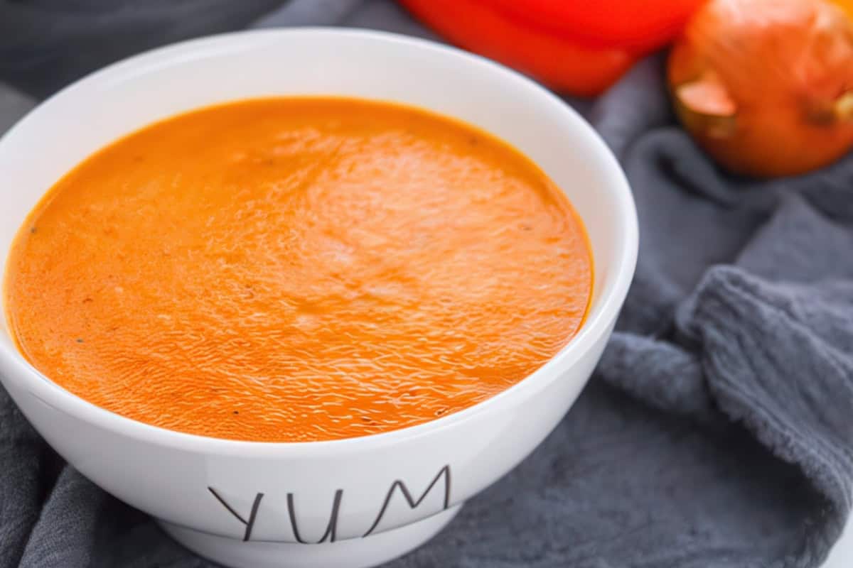 A white bowl labeled "YUM" is filled with creamy orange tomato soup. The bowl is placed on a gray cloth, with an onion and an orange cloth in the blurred background.