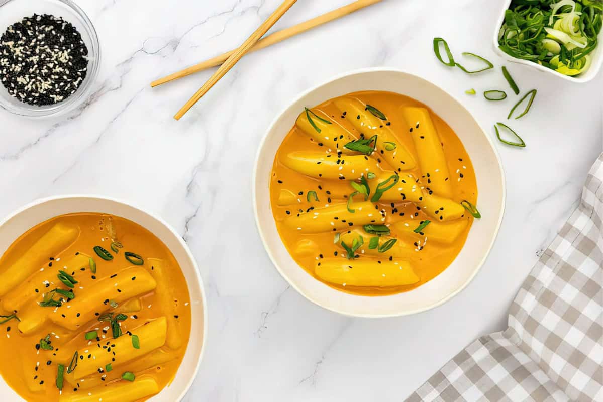 The image includes two bowls of Korean rice cakes in orange sauce, garnished with green onions and black sesame seeds. Nearby are chopsticks, a small bowl of black sesame seeds, and a dish of sliced green onions.