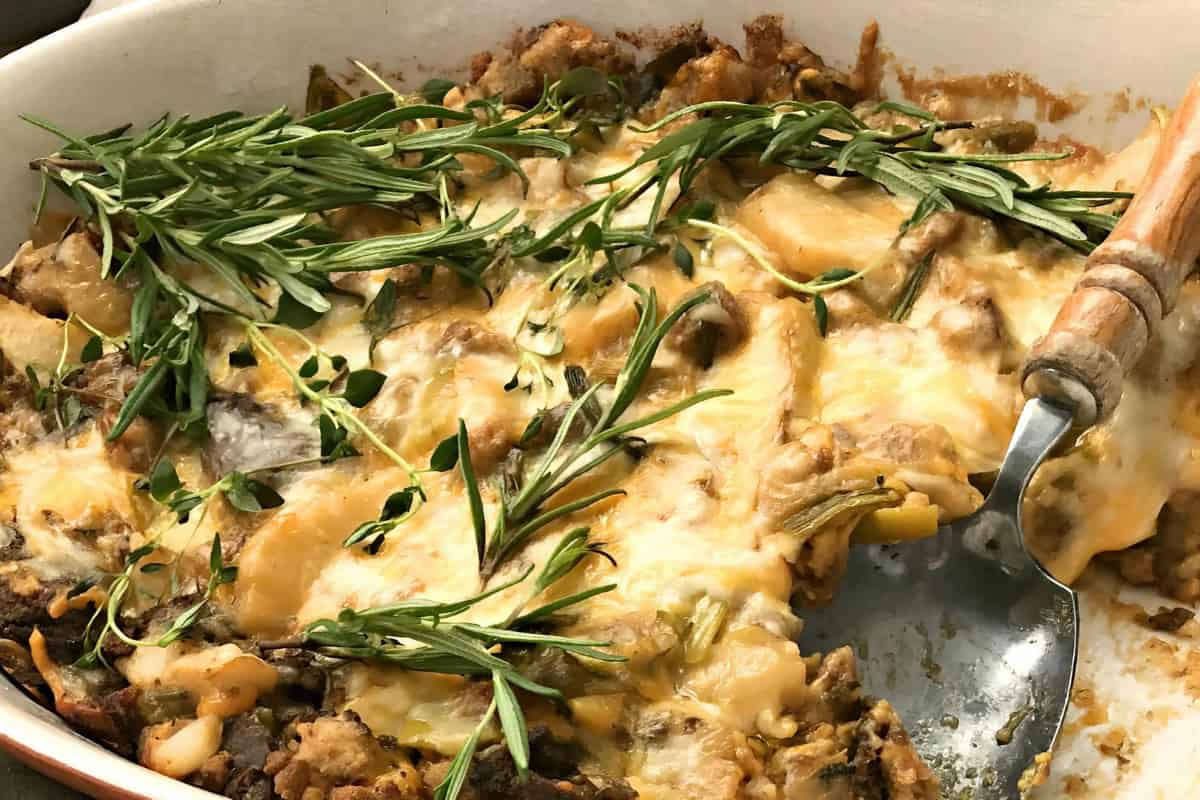 A baked casserole dish filled with melted cheese, cooked vegetables, and chunks of bread or meat. The dish is garnished with fresh rosemary and thyme. A serving spoon rests in the corner of the dish.