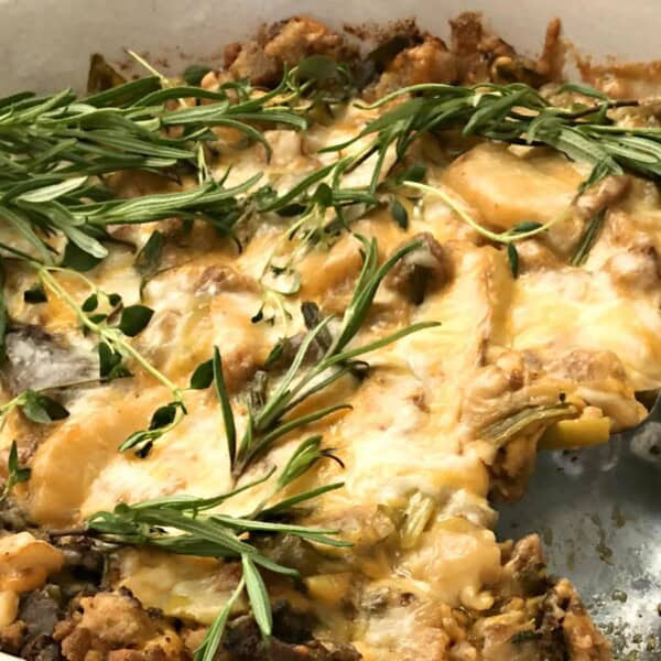 A casserole dish with a cheesy baked mix of vegetables and meat topped with rosemary, next to a serving spoon.