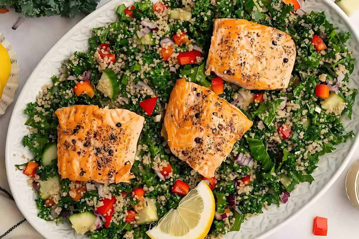 A plate of seasoned salmon fillets on a bed of couscous salad with kale, cucumber, bell peppers, and red onion. A lemon wedge is placed on the side for garnish.