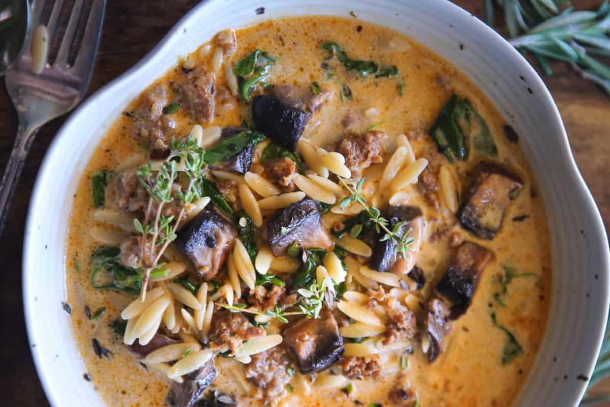 A bowl of creamy soup with orzo pasta, chunks of eggplant, ground sausage, and fresh spinach. The dish is garnished with sprigs of fresh herbs. A fork and a knife are partially visible on the wooden table beside the bowl.