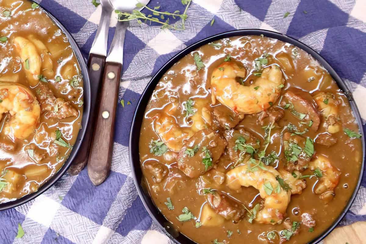 The image shows two bowls of shrimp and sausage gumbo garnished with herbs, set on a blue and white checkered cloth with forks nearby.