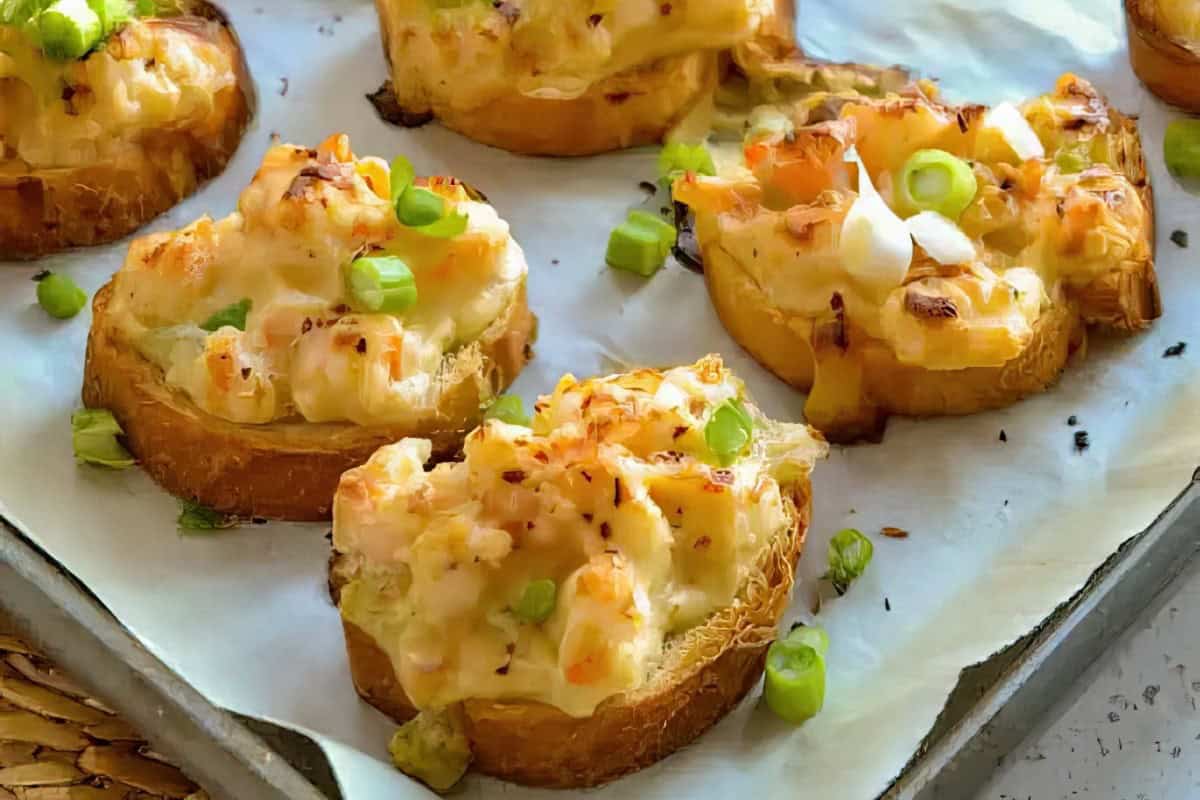Toasted bread slices topped with melted cheese, chopped shrimp, and green onions, arranged on a baking tray.