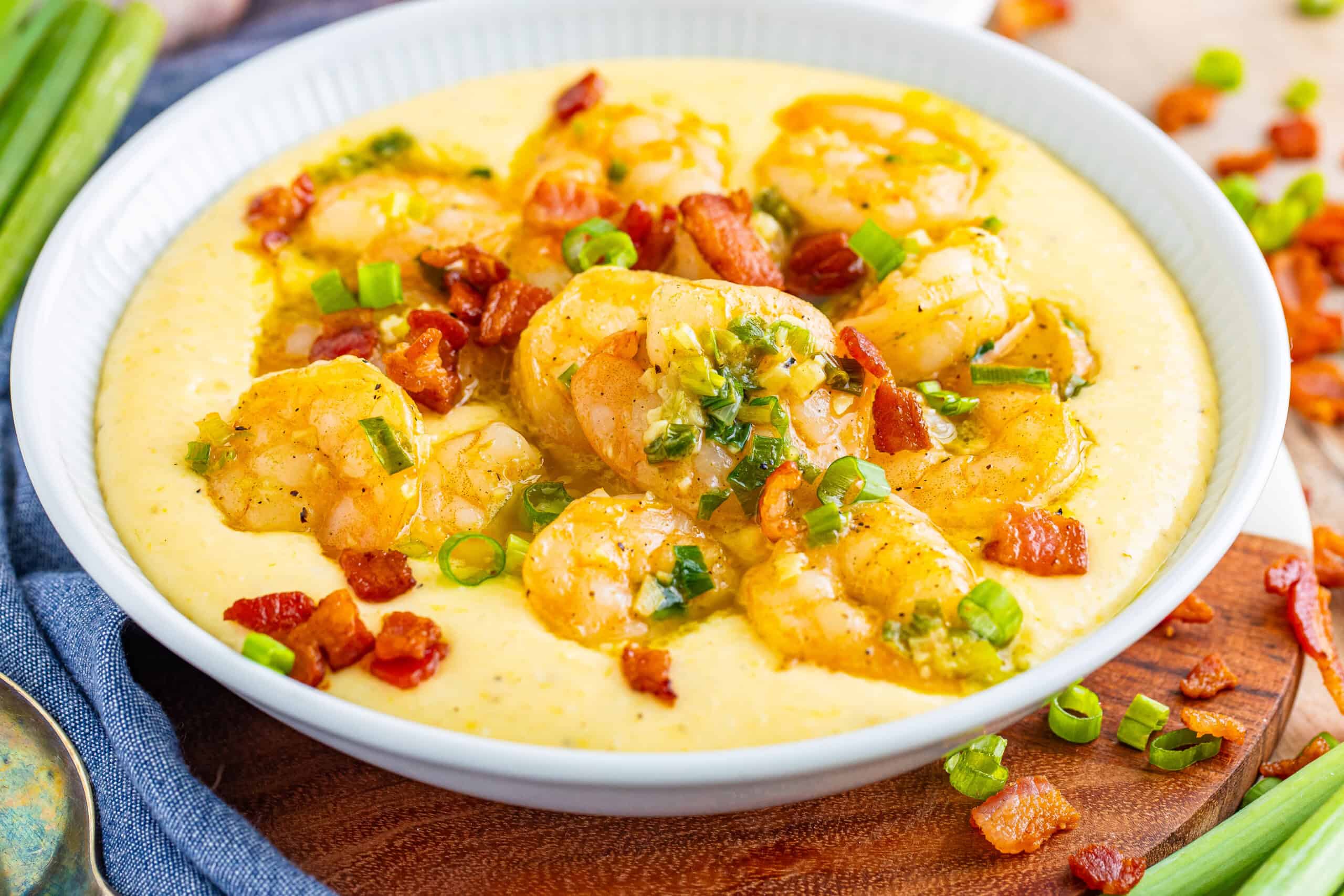 The image shows a bowl containing creamy grits, sautéed shrimp, crispy bacon bits, and chopped green onions.