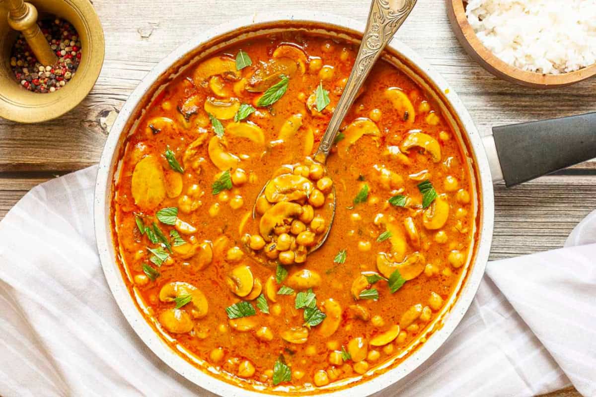 A pan filled with chickpea curry garnished with herbs, a spoon resting inside, rice, and a bowl of spices nearby.