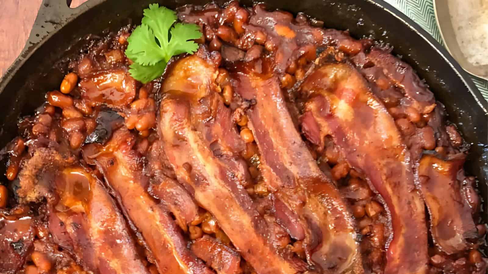 A skillet containing baked beans with strips of bacon, garnished with a sprig of cilantro.