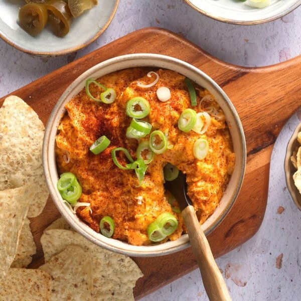 A bowl of creamy dip topped with green onions and paprika, on a wooden board with tortilla chips, hot sauce, and pickles.