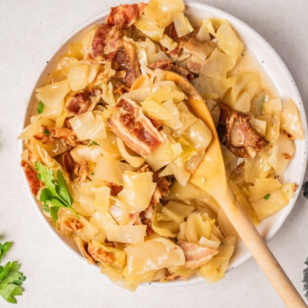 Cooked cabbage with bacon in a bowl, a wooden spoon on the side, fresh parsley, and a dark napkin nearby.