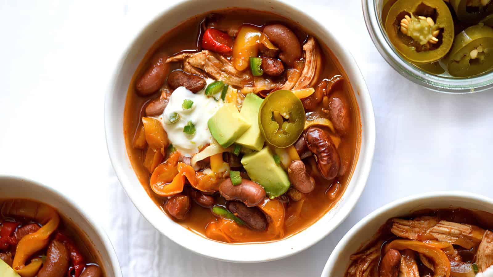 A bowl of chili with beans, shredded meat, and sliced peppers, topped with sour cream, avocado slices, and jalapeños. Additional bowls of chili are partially visible around it.
