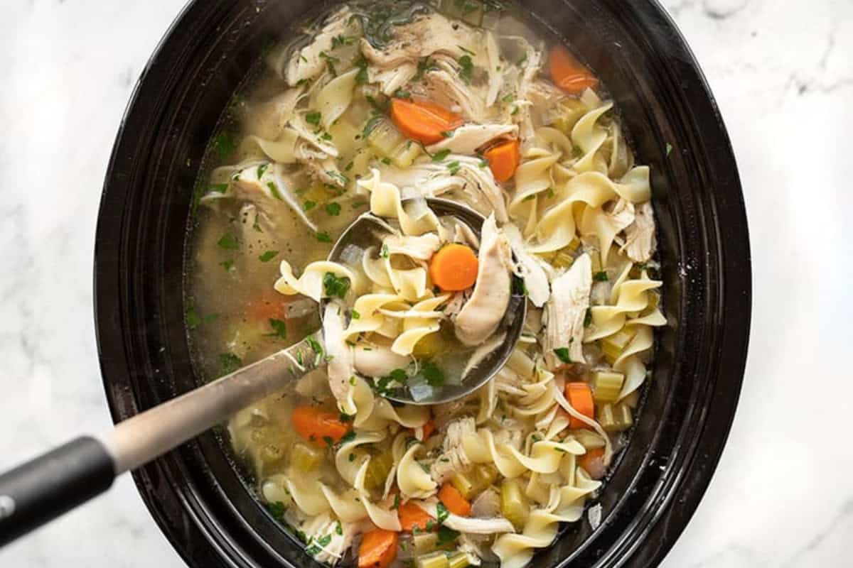 A ladle scoops hearty chicken noodle soup from a crockpot. The soup is filled with egg noodles, shredded chicken, sliced carrots, celery, and fresh parsley, all in a savory broth. The crockpot sits on a white marble surface.