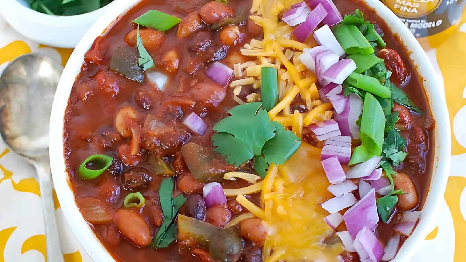 The image shows a bowl of chili garnished with shredded cheese, red onions, green onions, and cilantro. Next to the bowl are a spoon and a small dish containing green herbs.