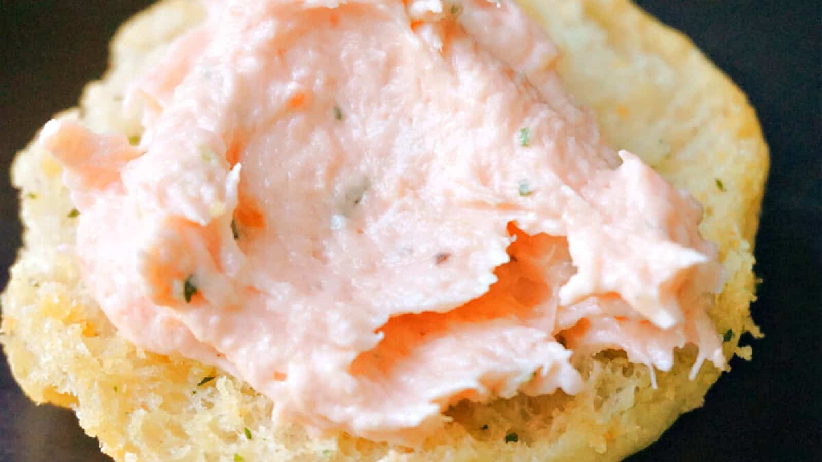 The image shows a close-up of a biscuit topped with a dollop of pink salmon spread and garnished with herbs.