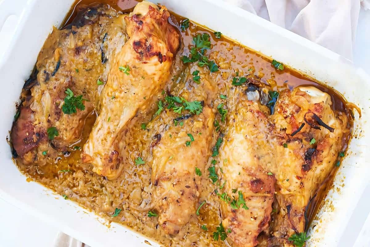 Baked chicken thighs and drumsticks in a creamy garlic sauce, garnished with fresh parsley, are displayed in a white baking dish on a light tablecloth. The dish has a golden, appetizing appearance.