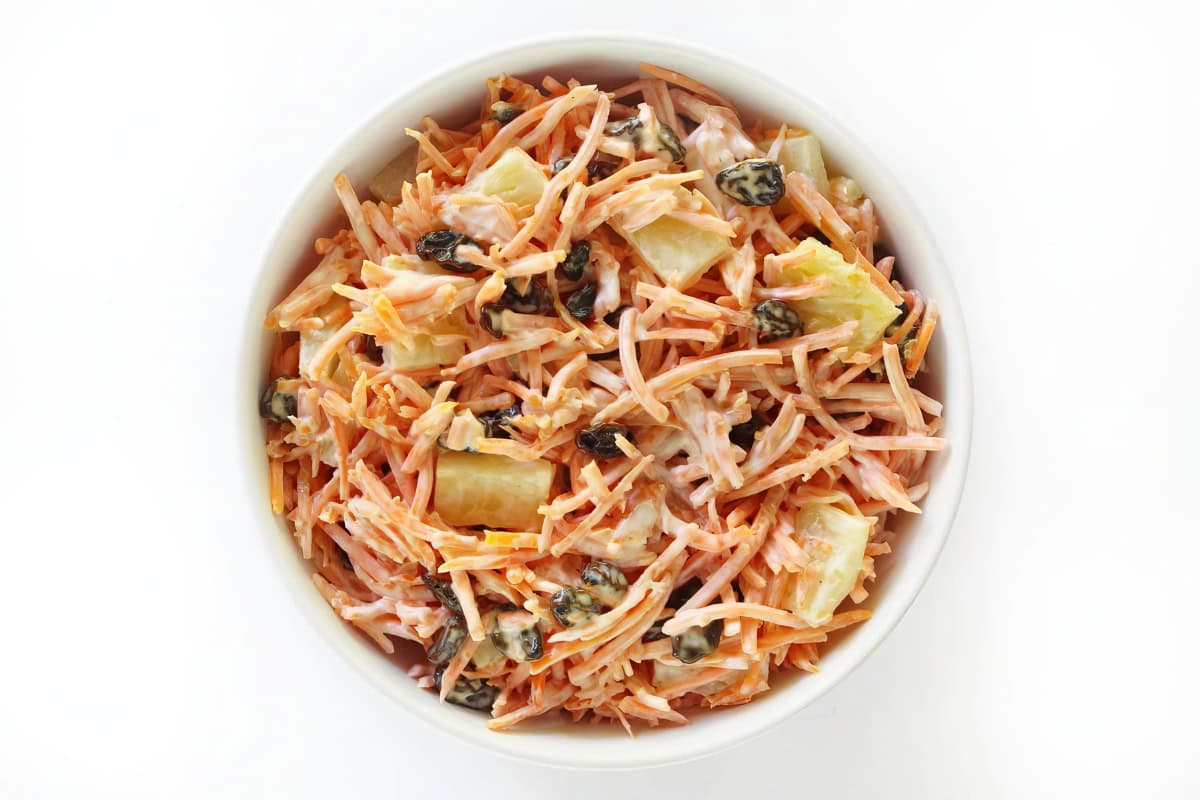 A bowl containing carrot salad, raisins, pineapple chunks, and creamy dressing.
