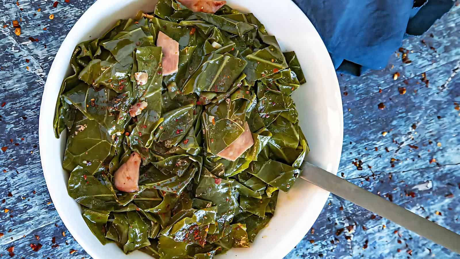 A bowl filled with cooked collard greens mixed with pieces of ham and spices, with a spoon resting inside. The bowl sits on a textured blue surface.