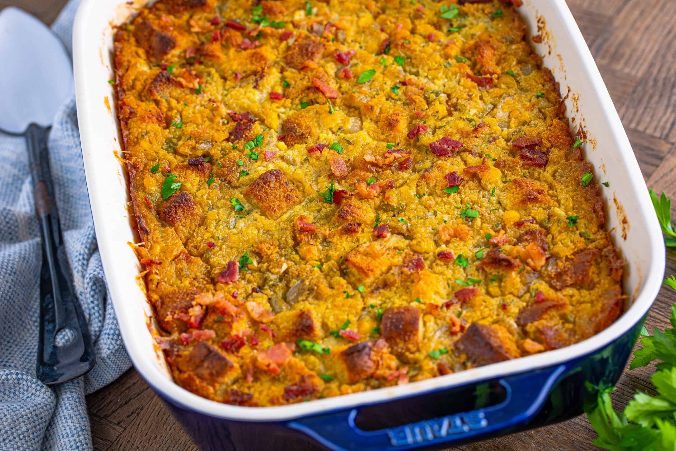 A blue dish filled with cornbread casserole, topped with diced bacon and herbs, resting on a wooden table.