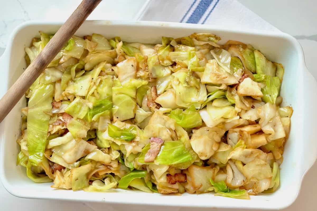 A white dish containing cooked cabbage and small bacon pieces, with a wooden spoon resting on the edge. A blue-striped cloth is partially visible in the background.