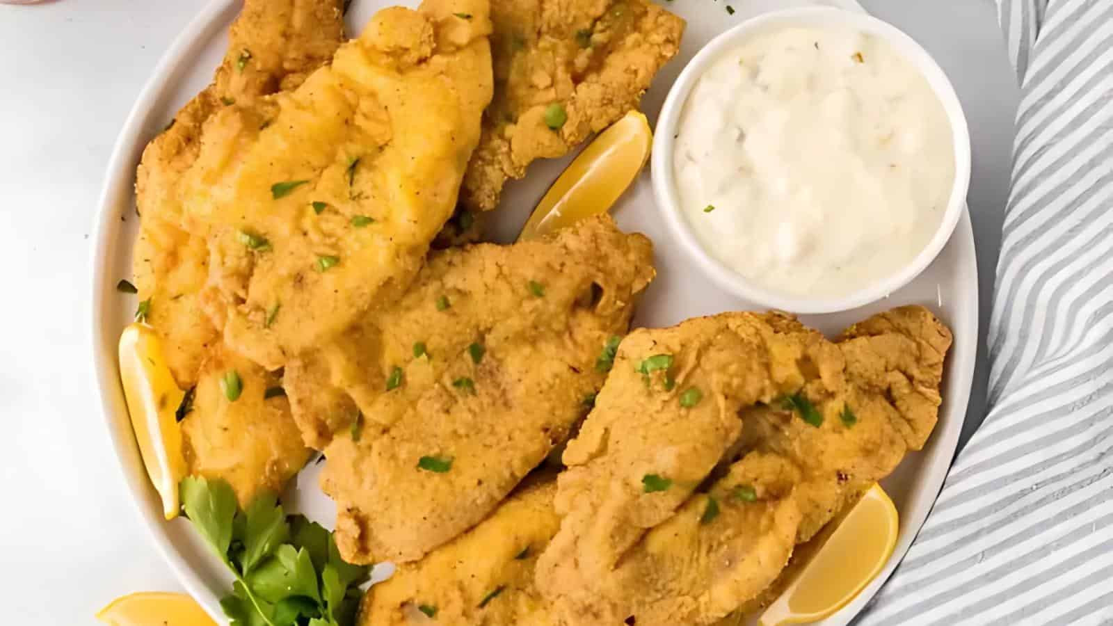 A plate of golden-brown fried fish fillets garnished with parsley, accompanied by lemon wedges and a bowl of creamy dipping sauce.