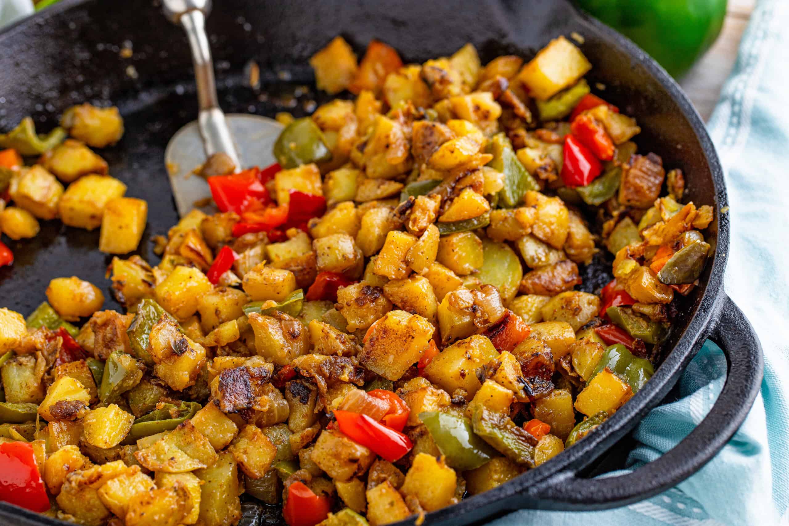 The image shows a skillet containing cooked diced potatoes, red and green peppers, and onions, with a metal spatula beside it.
