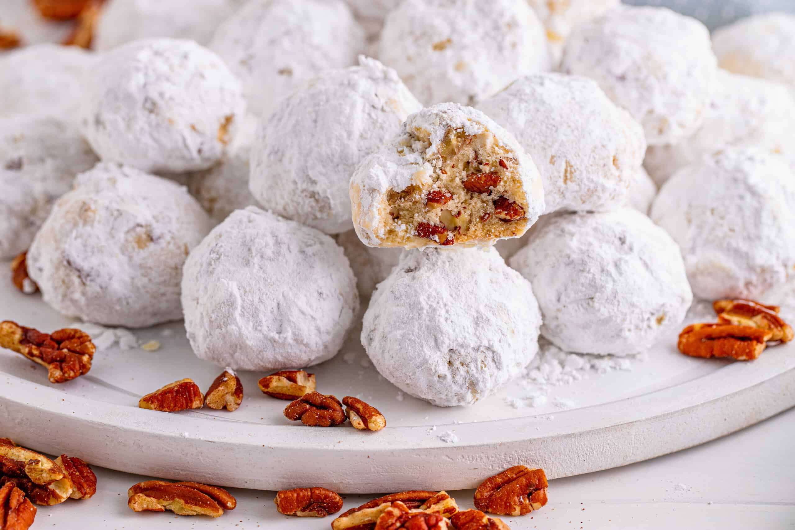 A plate of powdered sugar-coated cookies with exposed nutty interiors and scattered pecans surrounding the plate.