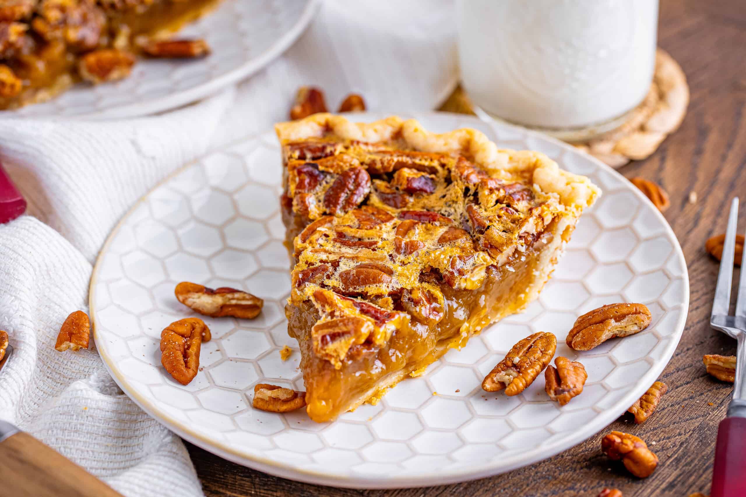 A slice of pecan pie on a white plate, a few pecans, and a glass of milk in the background.