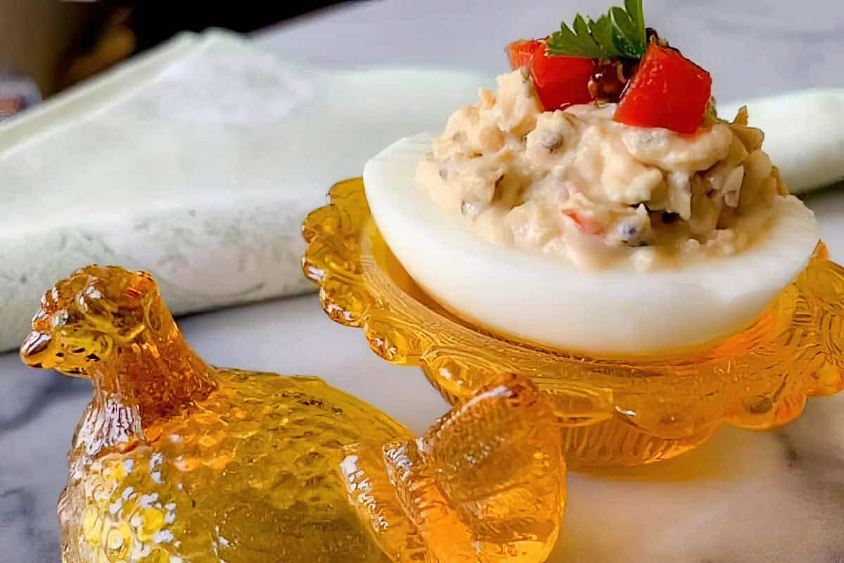 A deviled egg topped with red pepper and parsley, served in an ornate amber glass dish shaped like a chicken, placed on a marble surface with a white napkin in the background.