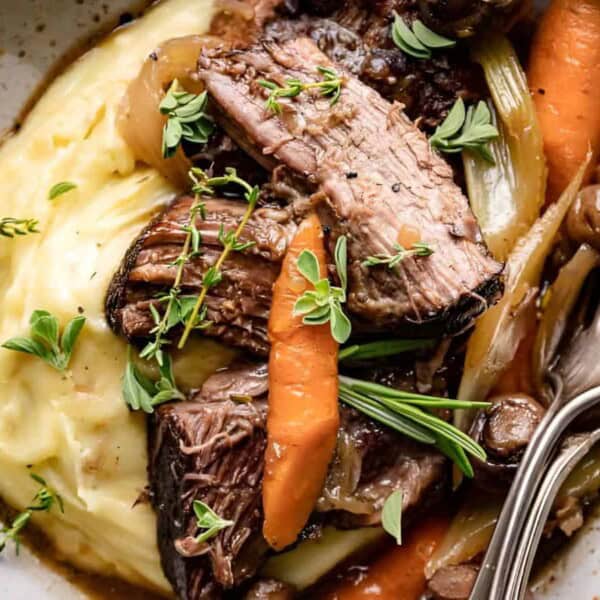 A plate of mashed potatoes topped with braised beef, carrots, mushrooms, fresh herbs, and a fork resting on the side.