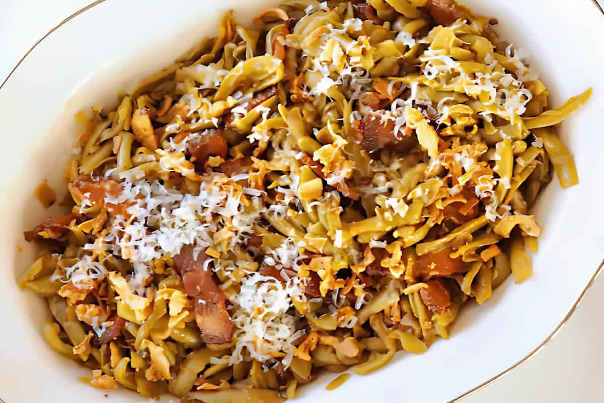 A close-up of a dish featuring fusilli pasta tossed with crispy bacon, mushrooms, and topped with grated Parmesan cheese. The pasta is served in a white bowl, highlighting the textures and colors of the ingredients.