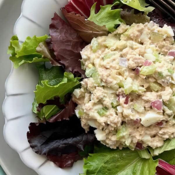 A white plate featuring a scoop of tuna salad on mixed greens with a fork beside it and a patterned cloth to the side.