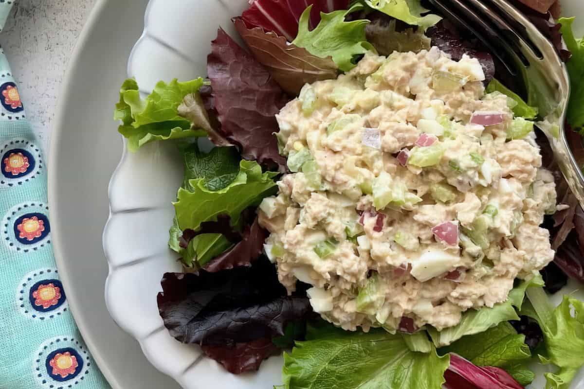 A white plate featuring a scoop of tuna salad on mixed greens with a fork beside it and a patterned cloth to the side.