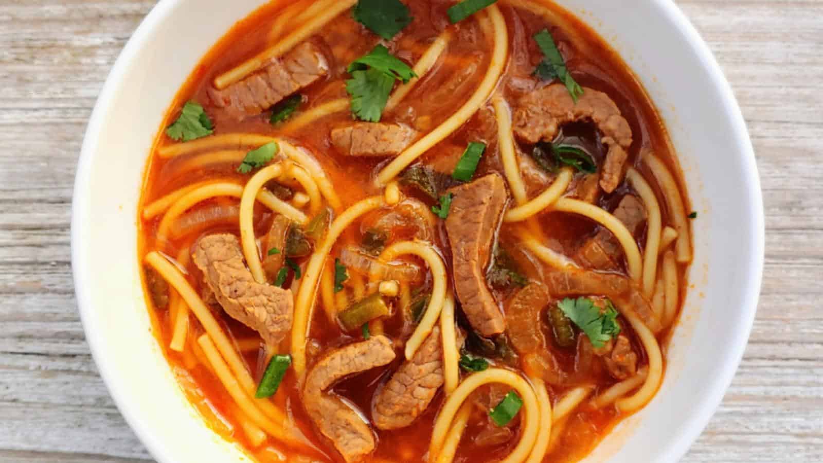 A bowl of spaghetti in rich tomato sauce, topped with slices of beef and garnished with fresh cilantro and chopped green onions. The dish is served on a light wooden table.