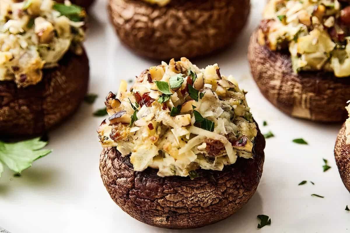 Close-up of stuffed mushrooms with a savory filling, garnished with chopped herbs and nuts on top. The filling appears creamy and is seasoned, sitting in large, cooked mushroom caps. A sprig of parsley is visible in the background.