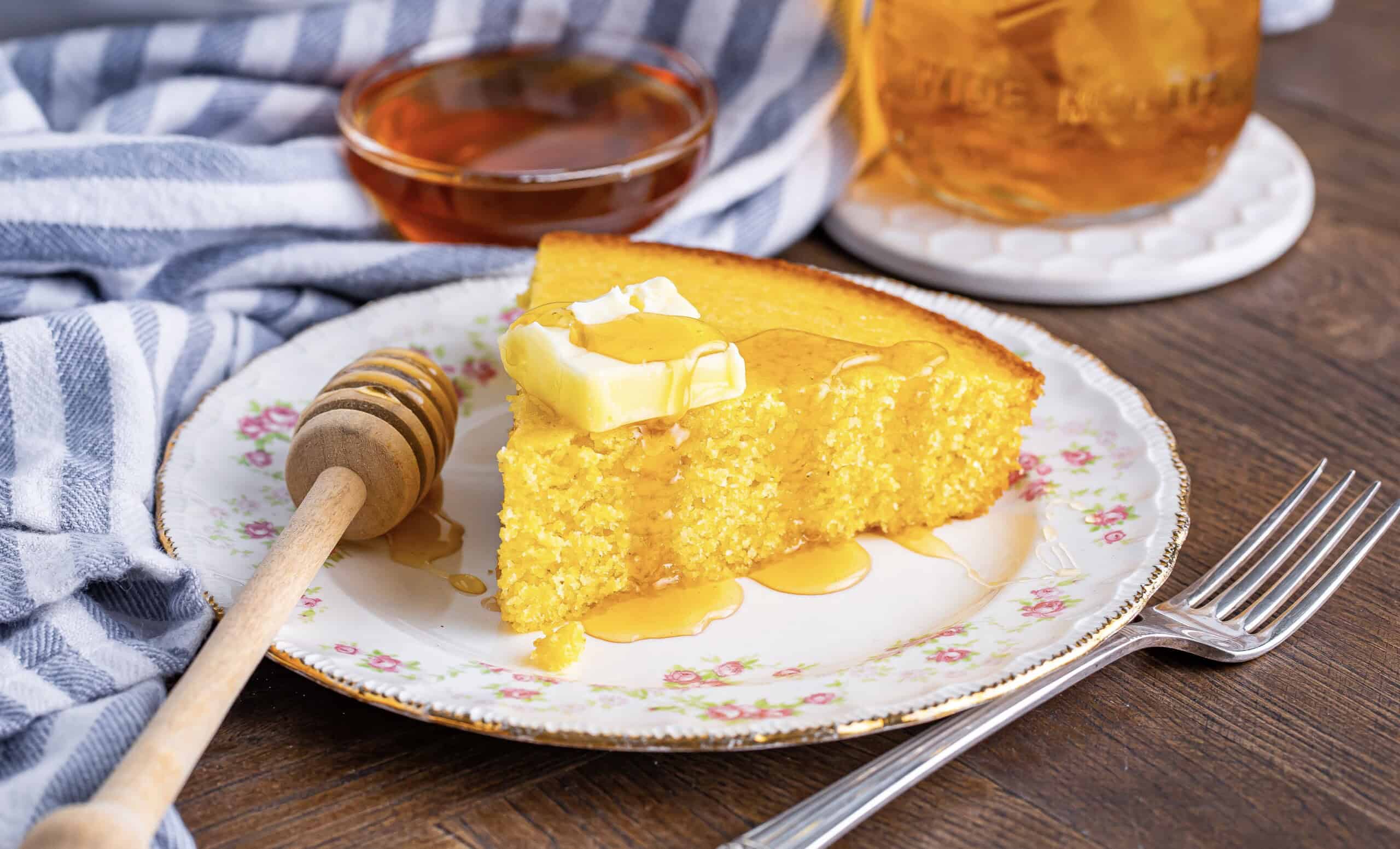 A slice of cornbread with butter and honey on a floral plate, accompanied by a honey dipper, a jar of amber liquid, and a striped cloth.