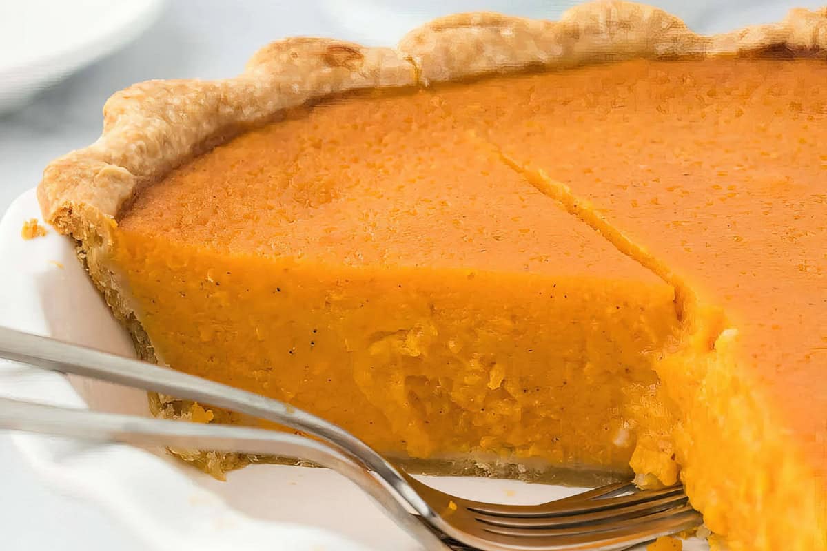 A sweet potato pie with a slice removed, revealing smooth orange filling and flaky crust, sits in a white pie dish with a serving utensil nearby.