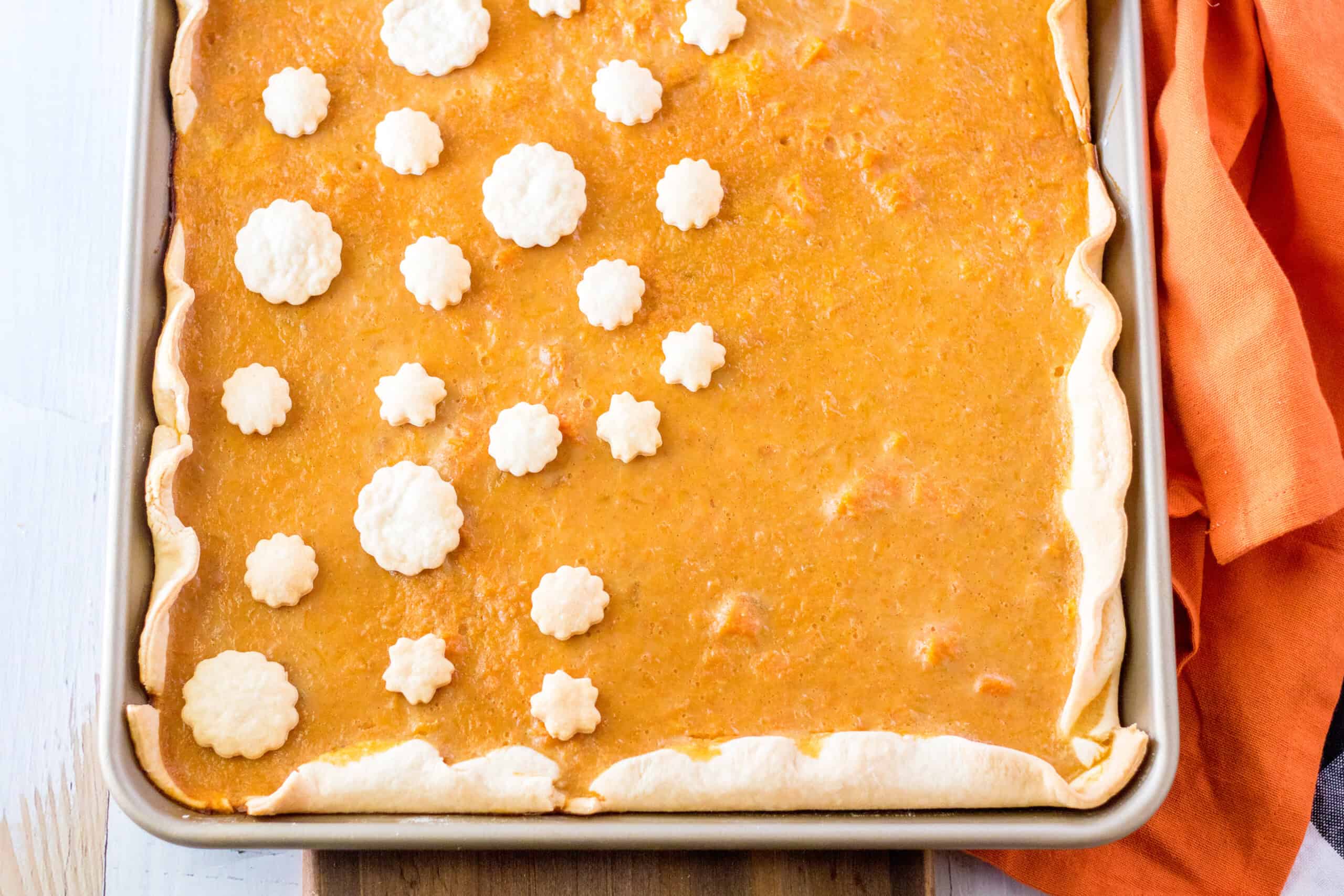 A rectangular pumpkin pie with a crimped edge crust in a baking pan, topped with small white pastry cutouts, placed beside an orange cloth.