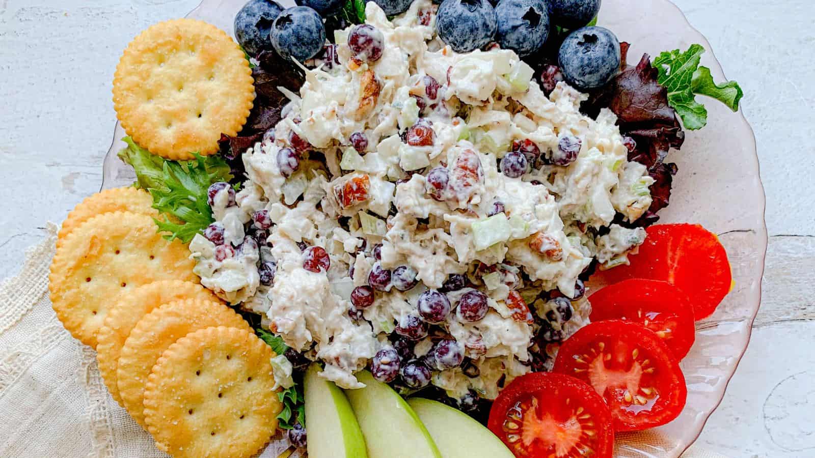 The image shows a plate with chicken salad as the main course. The salad includes grapes and nuts. Surrounding the plate are blueberries, crackers, lettuce, sliced tomatoes, and pear slices.