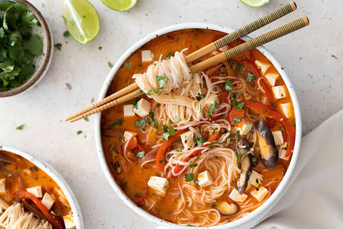 In the image, there is a bowl containing noodle soup along with tofu, mushrooms, and vegetables. The dish is garnished with herbs, and chopsticks are placed on top of the bowl. In the background, lime slices and cilantro are visible.